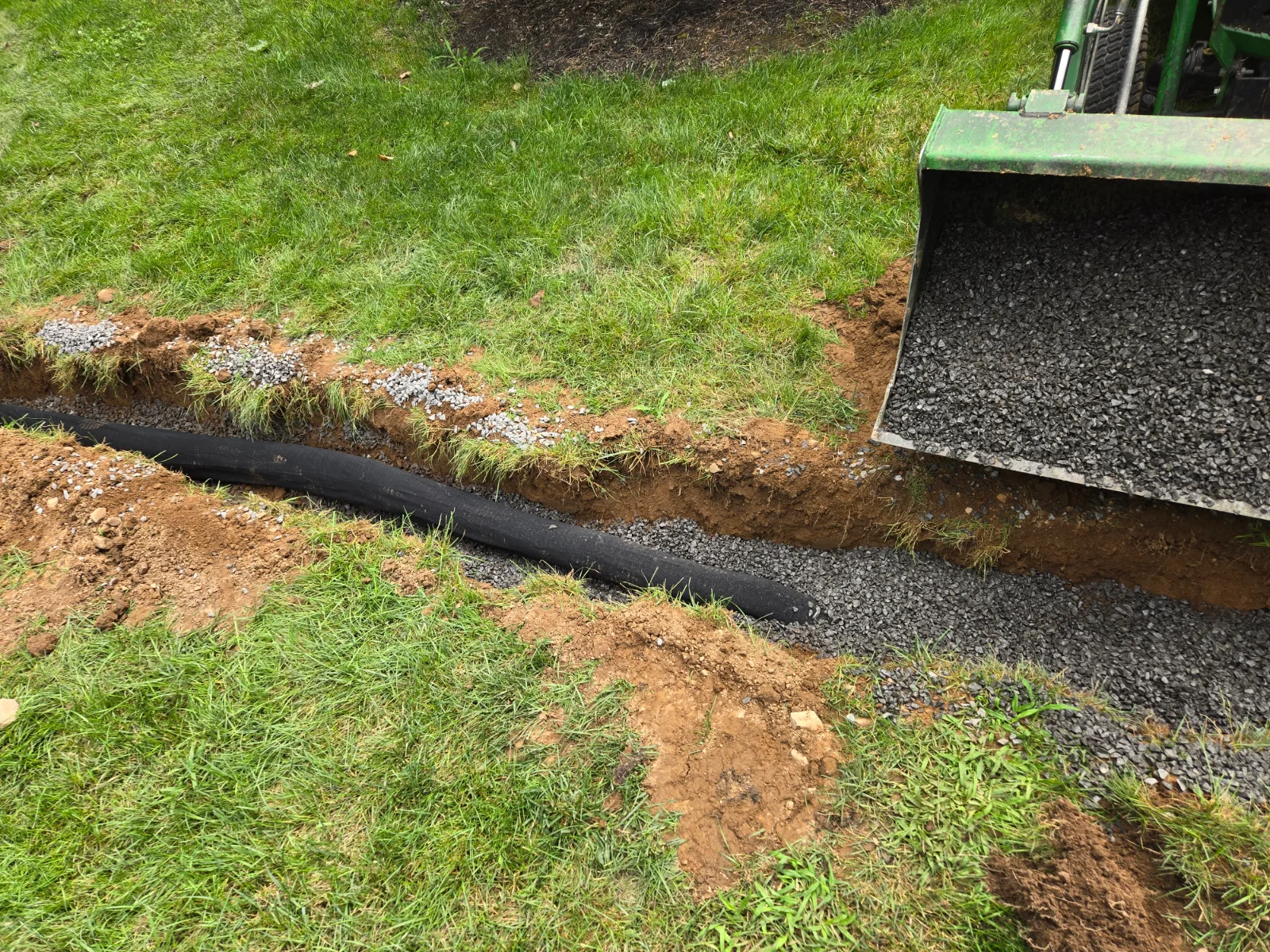 A tractor bucket of gravel beside a drainage trench in a lawn, with black drain pipe laid in it